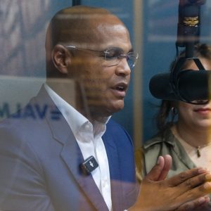 Marco Jensen hablando en un micrófono durante una sesión privada, vistiendo un traje azul y mostrando expresión concentrada detrás del cristal.